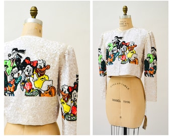 May include: A white sequined cropped jacket with a colorful embroidered design of Mickey Mouse and friends. The jacket has long sleeves and a button closure.