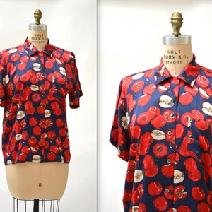 May include: A short-sleeved button-down shirt with a navy blue background and a red apple print. The shirt has a collar and a button-up front.