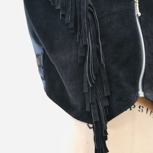May include: A black suede vest with a long fringe detail on the left side. The vest has a zipper closure.