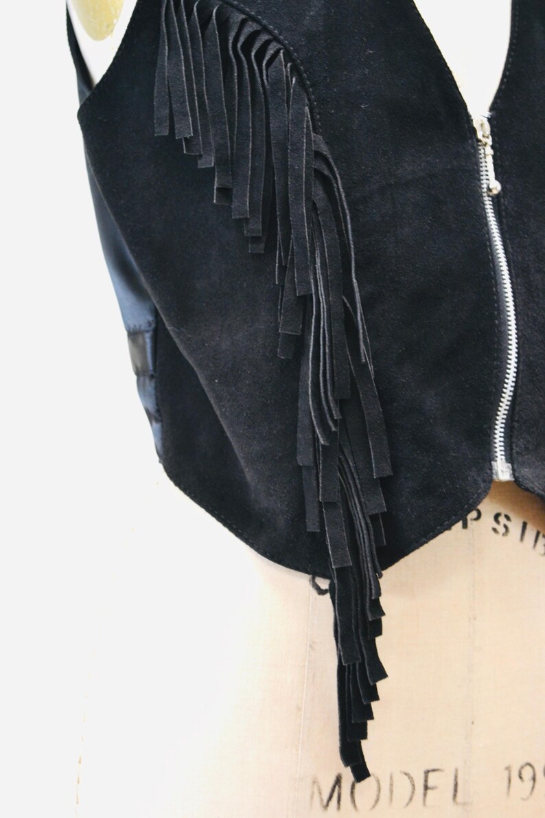 May include: A black suede vest with a long fringe detail on the left side. The vest has a zipper closure.