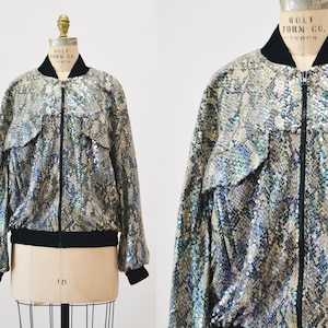 Pode incluir: Uma jaqueta bomber prateada iridescente com padrão de pele de cobra. A jaqueta tem um fecho de correr e dois bolsos.