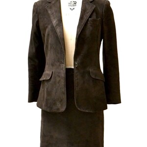 May include: A brown suede blazer and skirt suit. The blazer has a single button closure and two flap pockets. The skirt is knee-length.
