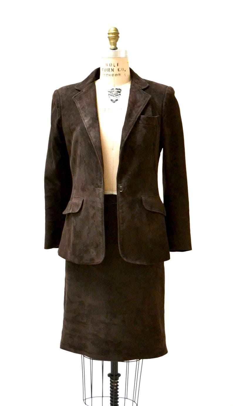 May include: A brown suede blazer and skirt suit. The blazer has a single button closure and two flap pockets. The skirt is knee-length.