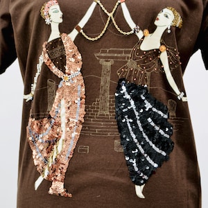 May include: A brown t-shirt with an illustration of two figures in classical dress, adorned with sequins. A gold chain connects the figures, set against a backdrop of architectural details.