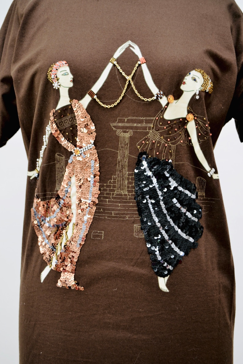 May include: A brown t-shirt with an illustration of two figures in classical dress, adorned with sequins. A gold chain connects the figures, set against a backdrop of architectural details.