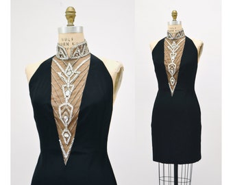 May include: A black halter dress with a high, embellished collar and a sheer panel down the front. The collar and panel feature intricate silver and clear crystal detailing. The dress is displayed on a mannequin.