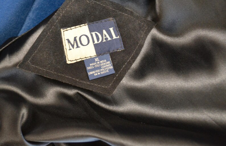May include: A black leather jacket with a blue lining. The label reads "MODAL" and indicates the jacket is size XL, made in India, with a shell of 100% genuine leather, lining of 100% polyester, and a  RN # 100374.