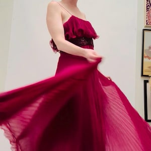 May include: A flowing, floor-length burgundy dress with a pleated skirt and spaghetti straps. The bodice features a ruffled design and a dark sequined band. The dress is being twirled, creating a dynamic and elegant look.