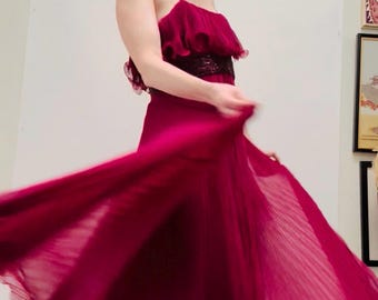 May include: A flowing, floor-length burgundy dress with a pleated skirt and spaghetti straps. The bodice features a ruffled design and a dark sequined band. The dress is being twirled, creating a dynamic and elegant look.