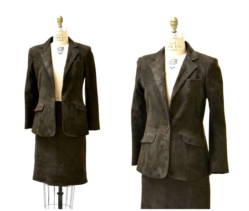 May include: A brown suede suit jacket and skirt set. The jacket has a single button closure and two pockets. The skirt is a-line and hits just above the knee.