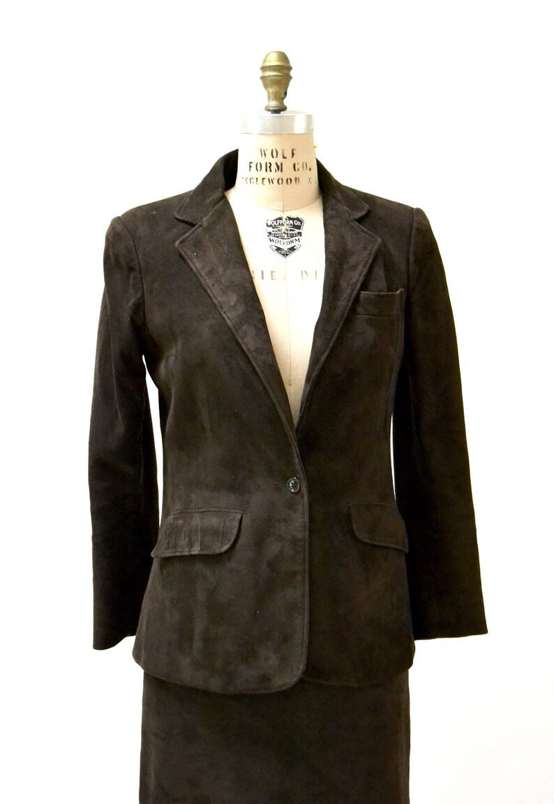 May include: A brown suede blazer and skirt suit. The blazer has a single button closure and two flap pockets. The skirt is knee-length.