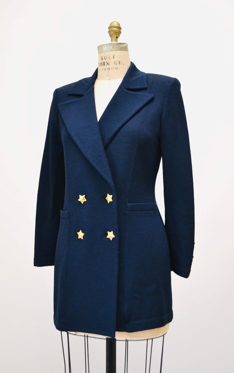 May include: A navy blue double-breasted blazer with gold star buttons. The blazer has a long, fitted silhouette and is made of a soft, textured fabric.