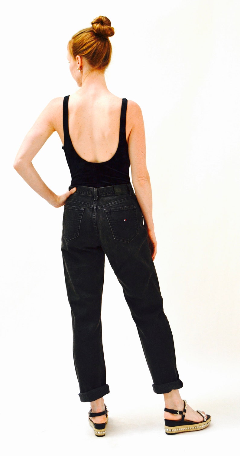 May include: A woman wearing a black tank top and black jeans with a small red and white logo on the back pocket. She is wearing black sandals with a gold platform.