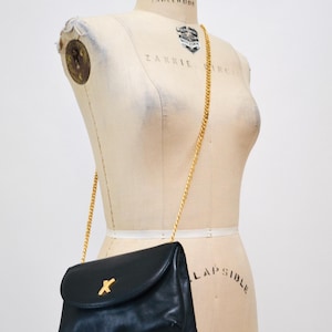 May include: A black leather shoulder bag with a gold chain strap and a gold X-shaped closure. The bag is on a white dress form.