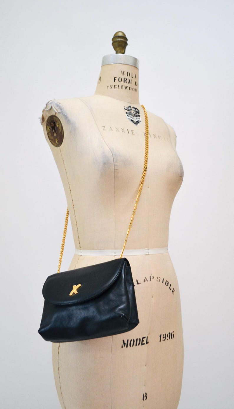 May include: A black leather shoulder bag with a gold chain strap and a gold X-shaped closure. The bag is on a white dress form.