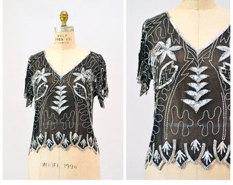 Puede incluir: Una blusa negra transparente con lentejuelas y bordados plateados. La blusa tiene un escote en V y mangas cortas. El bordado presenta un diseño floral con hojas y flores. La blusa está etiquetada como "MODFL 1994".