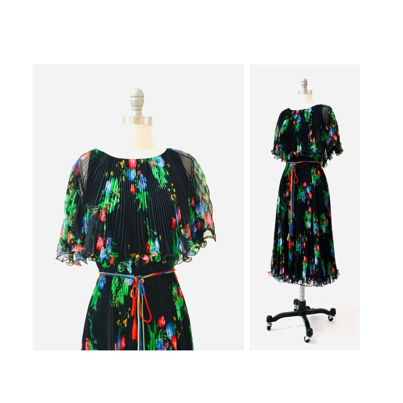 May include: A black pleated dress with a floral print and a matching cape. The dress has a tie at the waist and is made of a sheer fabric.