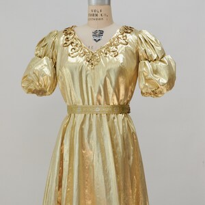May include: A gold metallic dress with a V-neckline and puff sleeves. The dress has a beaded embellishment around the neckline and a thin gold belt at the waist.