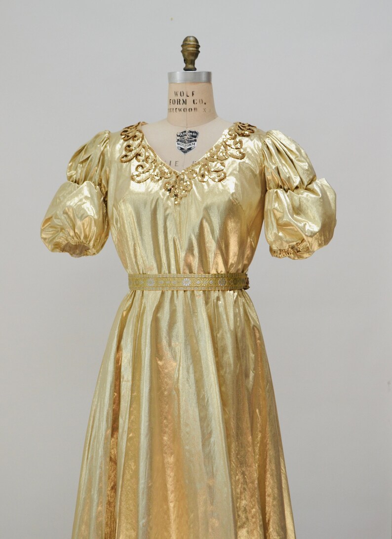 May include: A gold metallic dress with a V-neckline and puff sleeves. The dress has a beaded embellishment around the neckline and a thin gold belt at the waist.