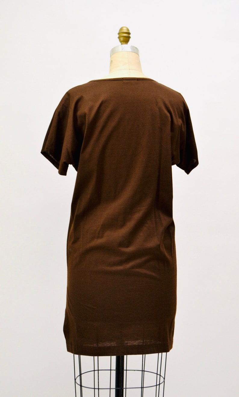 May include: A brown t-shirt dress with short sleeves, shown on a mannequin. The dress features a scoop neckline and a straight, relaxed fit. The fabric looks soft and solid-coloured, ideal for casual wear. The dress is a medium length.