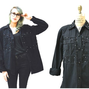 May include: Black denim shirt with long sleeves, decorated with small, silver studs. The shirt has two chest pockets and a classic collar. The cuffs are rolled up. The shirt is buttoned up and has a relaxed fit. The shirt is on a mannequin.