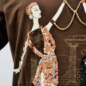 May include: A brown long-sleeved top with an art deco design. The design features a woman in a sequined dress, with gold chain accents and a beaded necklace. The background includes architectural elements in a sepia tone.