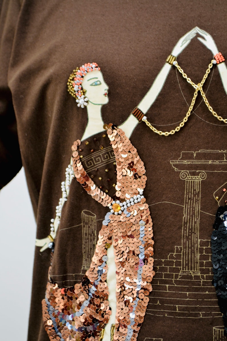 May include: A brown long-sleeved top with an art deco design. The design features a woman in a sequined dress, with gold chain accents and a beaded necklace. The background includes architectural elements in a sepia tone.