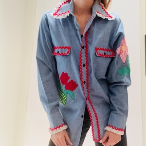 Puede incluir: Camisa vaquera azul claro con ribetes rojos y blancos. La camisa presenta aplicaciones de tulipanes y flores rojas. El cuello y los puños tienen ribetes de encaje blanco. La camisa tiene dos bolsillos y una botonadura.
