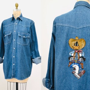 May include: A blue denim shirt with a long sleeve and button-down front. The shirt has two chest pockets and a cartoon character embroidered on the back. The character is a duck wearing a cowboy hat and holding a gun.