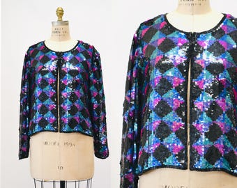 May include: A black sequined jacket with a geometric pattern in shades of blue, pink, and purple. The jacket has a front zipper closure and long sleeves.
