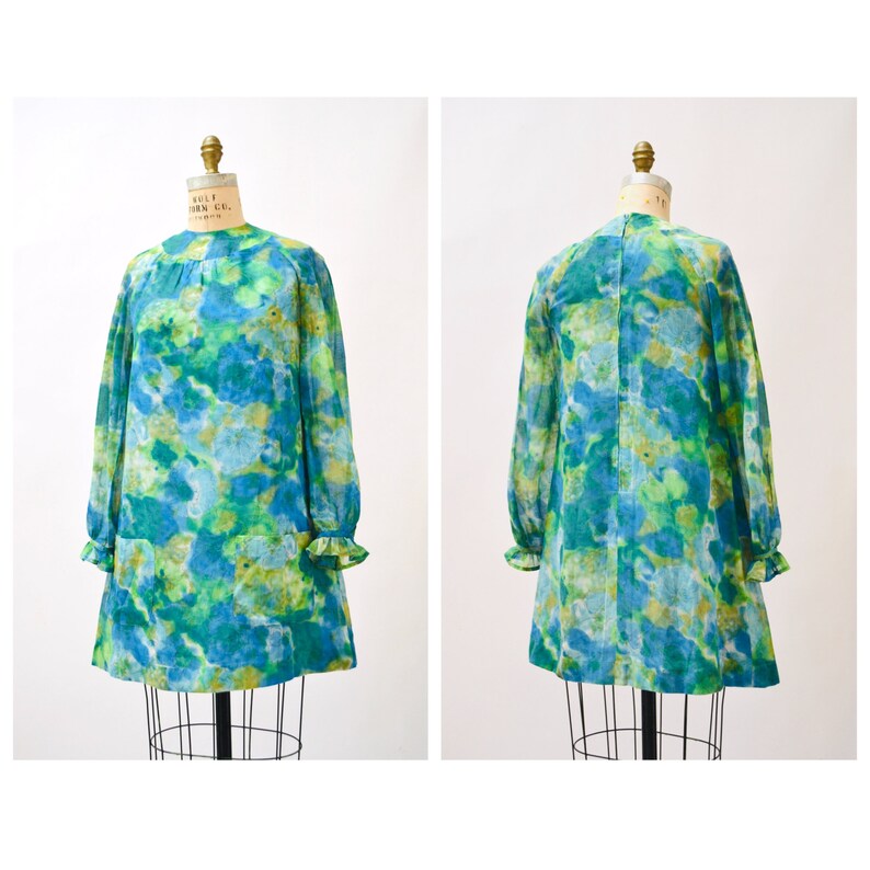 May include: A vintage green and blue floral print mini dress with long sleeves and a round neckline. The dress has a loose fit and a slightly flared skirt.