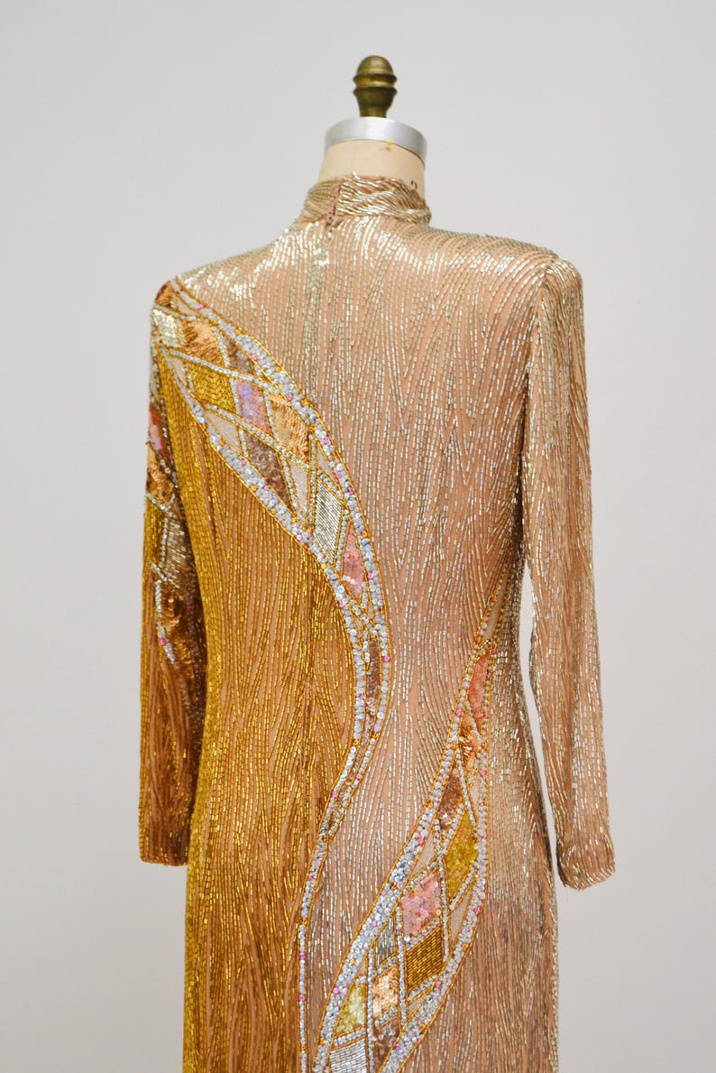 Op de afbeelding: Een lange, met gouden pailletten bezette jurk met een geometrisch patroon van iriserende pailletten. De jurk heeft een hoge halslijn en lange mouwen.