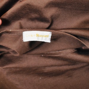 May include: Close-up of brown fabric with a tag reading "Didier Ronsard" in gold lettering. The fabric is folded, possibly a garment. The tag is white with a gold border.