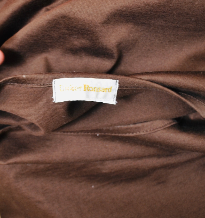 May include: Close-up of brown fabric with a tag reading "Didier Ronsard" in gold lettering. The fabric is folded, possibly a garment. The tag is white with a gold border.