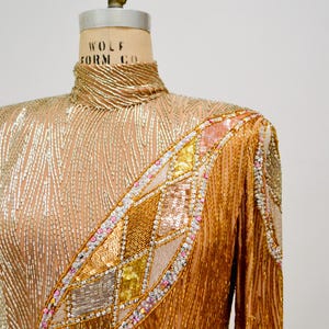 Op de afbeelding: Een lang mouw, hoog gesloten, gouden pailletten top met een geometrisch patroon van pailletten in tinten roze, goud en zilver. De top is op een paspop.
