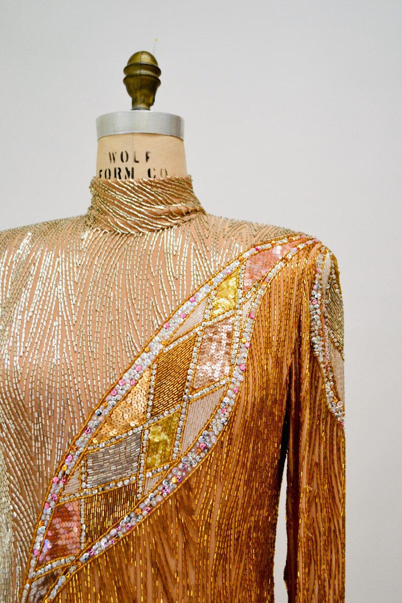 Op de afbeelding: Een lang mouw, hoog gesloten, gouden pailletten top met een geometrisch patroon van pailletten in tinten roze, goud en zilver. De top is op een paspop.