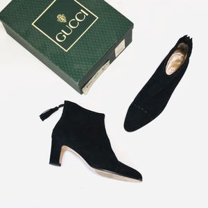 May include: Black suede ankle boots with a pointed toe and a small heel. The boots have a zipper closure and a tassel detail. A green Gucci box is in the background.