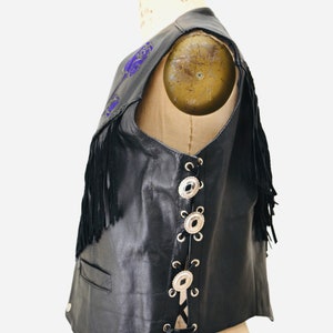 K&ouml;nnte beinhalten: Schwarze Lederweste mit Fransen und silbernen Concho-Kn&ouml;pfen. Die Weste hat einen Schn&uuml;rr&uuml;cken und violette Stickereien.