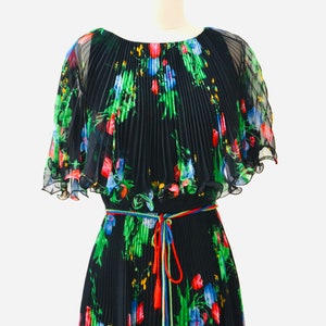 May include: A black pleated maxi dress with a floral print in shades of red, blue, green, and yellow. The dress has a cape-like top and a tie belt with tassels.