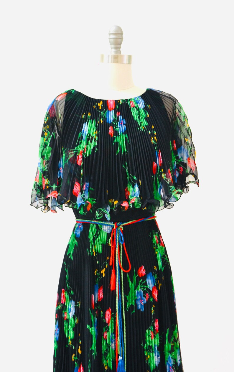 May include: A black pleated maxi dress with a floral print in shades of red, blue, green, and yellow. The dress has a cape-like top and a tie belt with tassels.