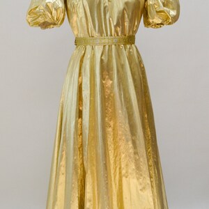 May include: A gold metallic dress with a gathered neckline and puff sleeves. The dress has a fitted bodice and a flowing skirt. The dress is cinched at the waist with a gold and white ribbon.