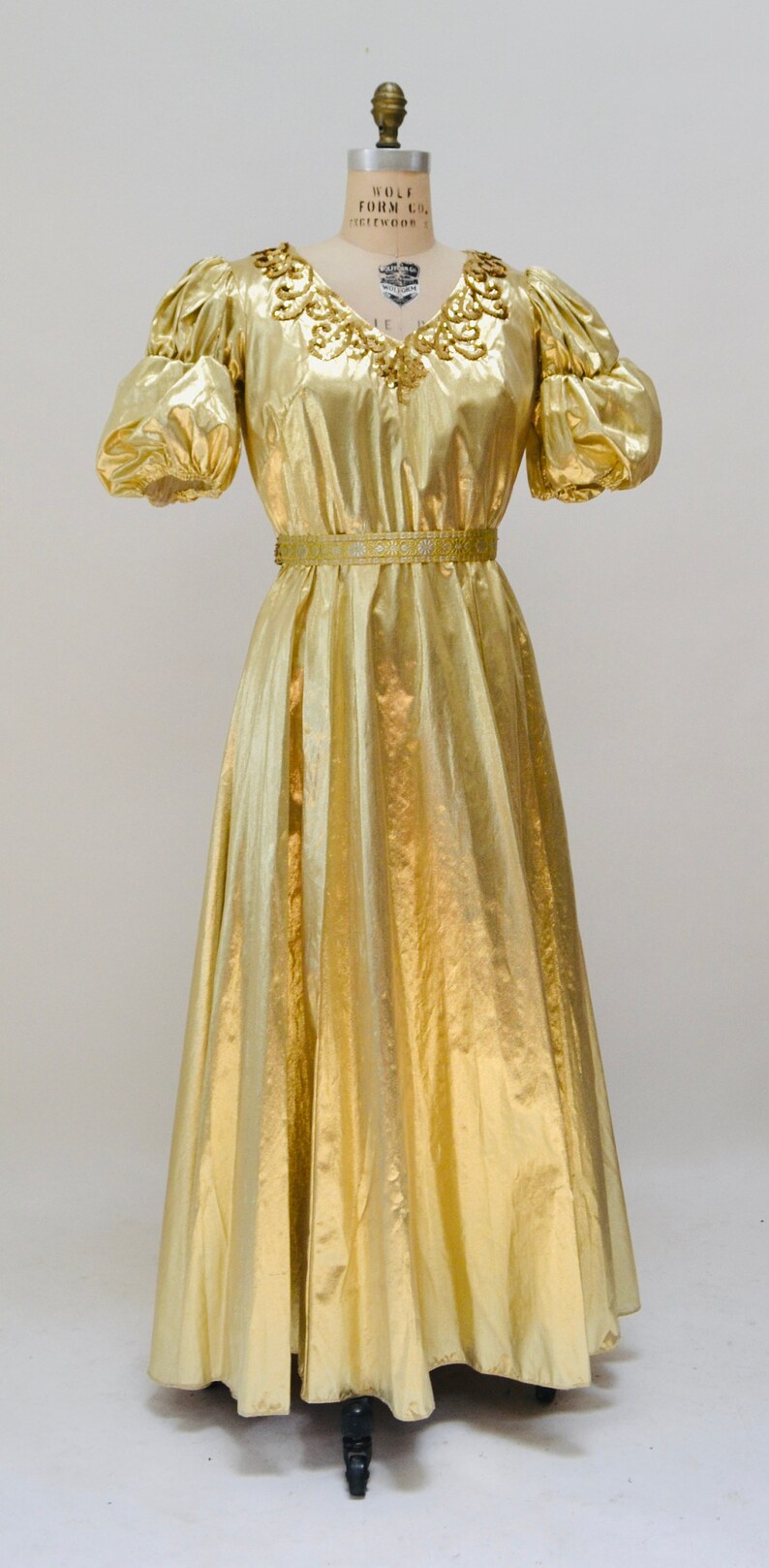 May include: A gold metallic dress with a gathered neckline and puff sleeves. The dress has a fitted bodice and a flowing skirt. The dress is cinched at the waist with a gold and white ribbon.