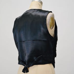 May include: A black satin vest with a fringed trim. The vest has a tie closure at the back.