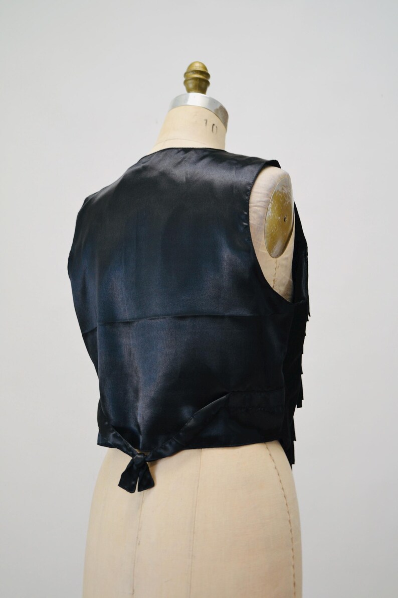 May include: A black satin vest with a fringed trim. The vest has a tie closure at the back.