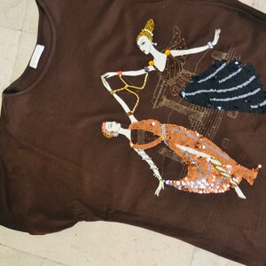 May include: A brown t-shirt with an Art Deco design. Two stylized female figures are embellished with black, copper, and gold sequins. The design incorporates architectural elements.