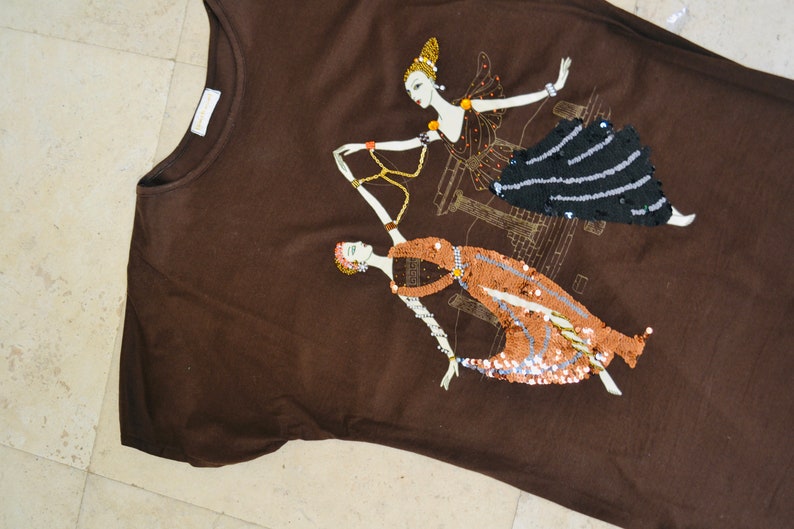 May include: A brown t-shirt with an Art Deco design. Two stylized female figures are embellished with black, copper, and gold sequins. The design incorporates architectural elements.