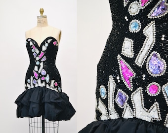May include: A black mini dress with a strapless sweetheart neckline. The dress is covered in sequins and colorful geometric shapes. The bottom of the dress has a ruffled skirt.
