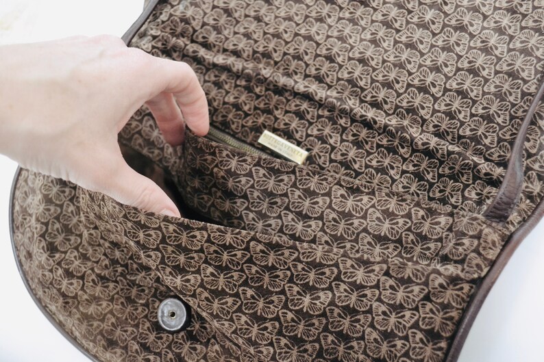 May include: The inside of a brown leather bag with a butterfly print lining. The bag has a zipper closure and a small metal tag that reads "Nine West".