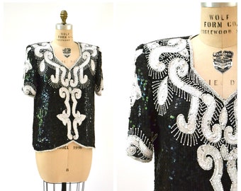 Puede incluir: Una blusa negra de lentejuelas con adornos de cuentas plateadas y blancas. La blusa tiene cuello en V y mangas cortas.