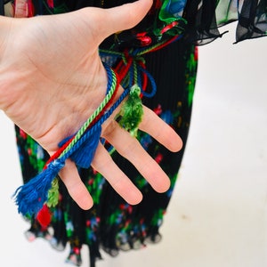 May include: Black pleated maxi dress with a floral print and colorful tassel ties. The dress has a fitted bodice and a flowing skirt.